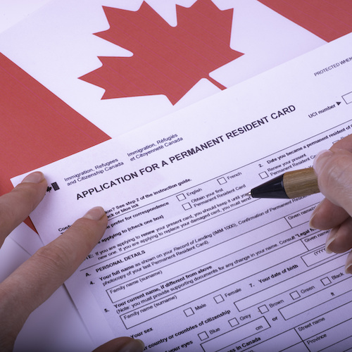 Filling Application (female hands and wood pen) form for Canadian Citizenship - Adults. Immigration, Refugees and Citizenship Canada on Canadian flag surface.